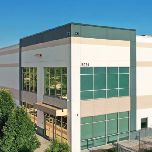 A modern industrial building with a large green facade and multiple windows sits on a paved area near trees A chainlink